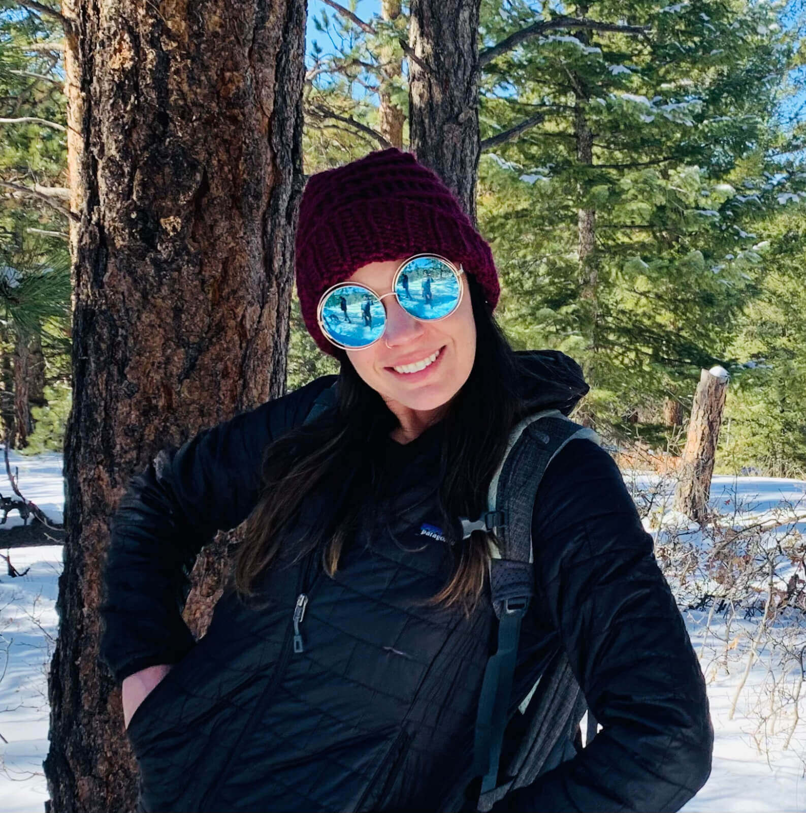 A woman leans against a tree wearing a coat, hat and circular mirror sunglasses in the forest carpeted with snow.