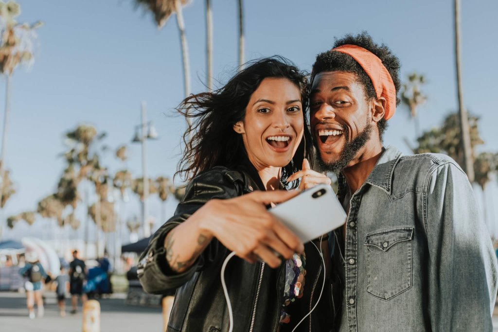 Couple smiling and taking a selfie in a sunny place with palm tree in the background.