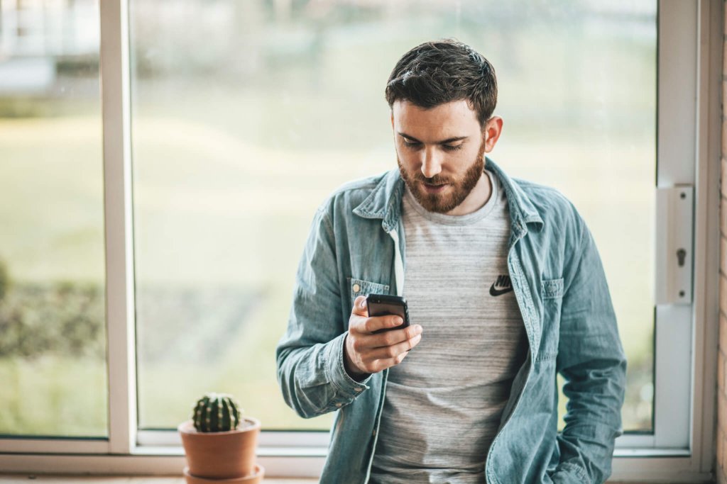 Let Us Help Showcase Your Social Media Content! Man standing in front of window looking at his smartphone.